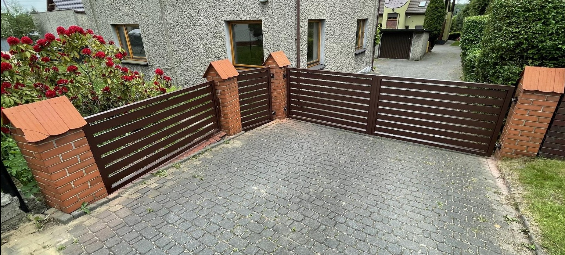 Automatyczna brama wjazdowa z poziomymi panelami, osadzona na ceglanych słupkach z daszkami, na tle domu z szarą elewacją i brukowanego podjazdu.