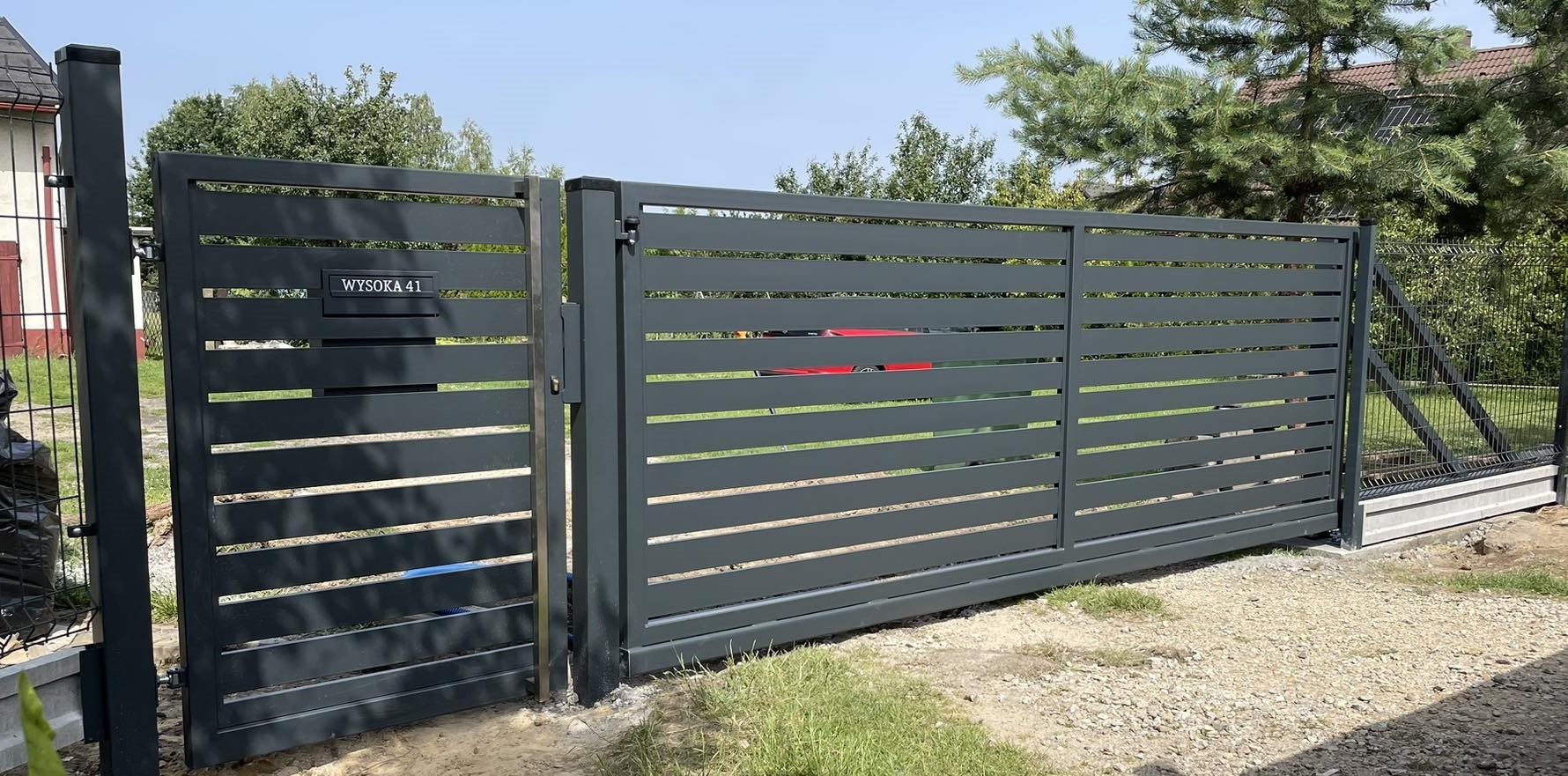 Nowoczesna, przesuwna brama wjazdowa w kolorze grafitowym z poziomymi panelami, z numerem domu 'Wysoka 41', częściowo otwarta, widoczny fragment ogrodzenia panelowego w tle.