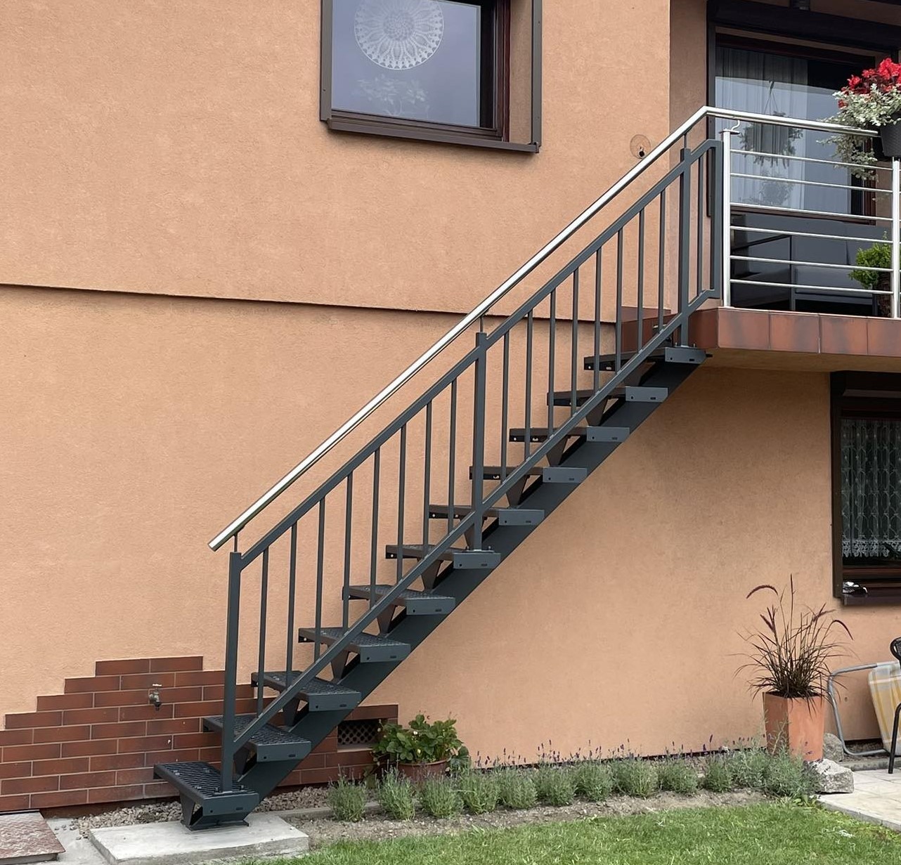 Zewnętrzne schody metalowe z poręczą ze stali nierdzewnej prowadzące na balkon, przymocowane do elewacji budynku z dekoracyjnym oknem i roślinami w donicach.