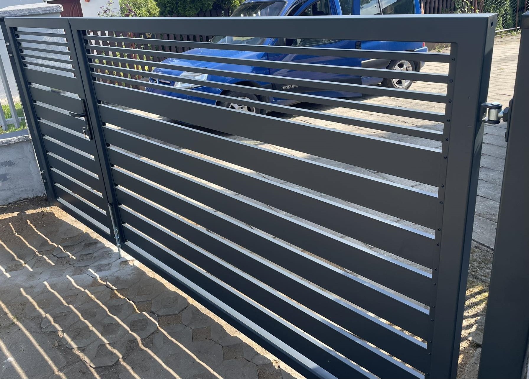 Nowoczesna, antracytowa brama z poziomymi panelami, widoczny mechanizm otwierania i częściowo przysłonięty samochód w tle.