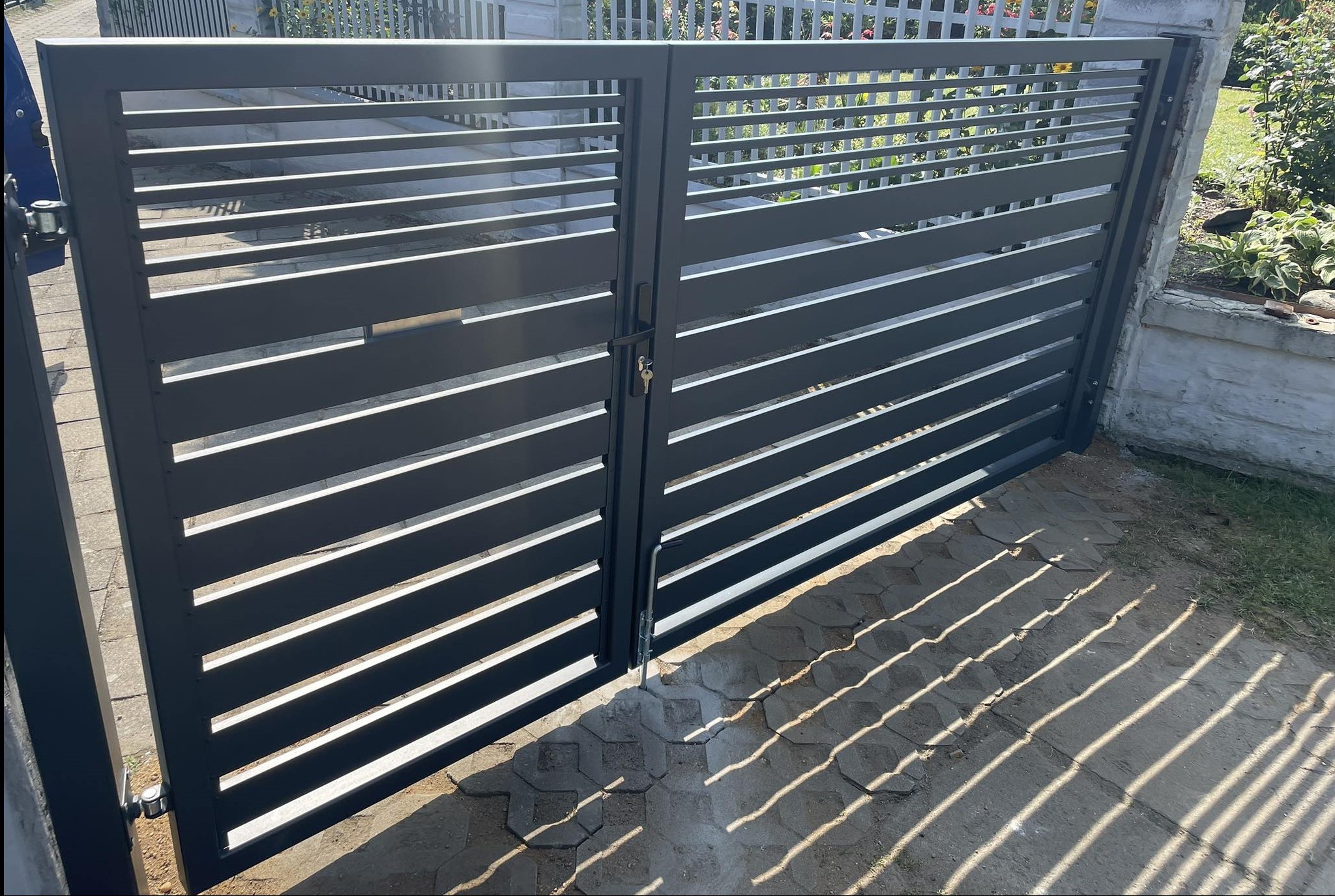Nowoczesna, dwuskrzydłowa, metalowa brama z poziomymi profilami, widoczne klucze w zamku, cień rzucany na kostkę brukową.