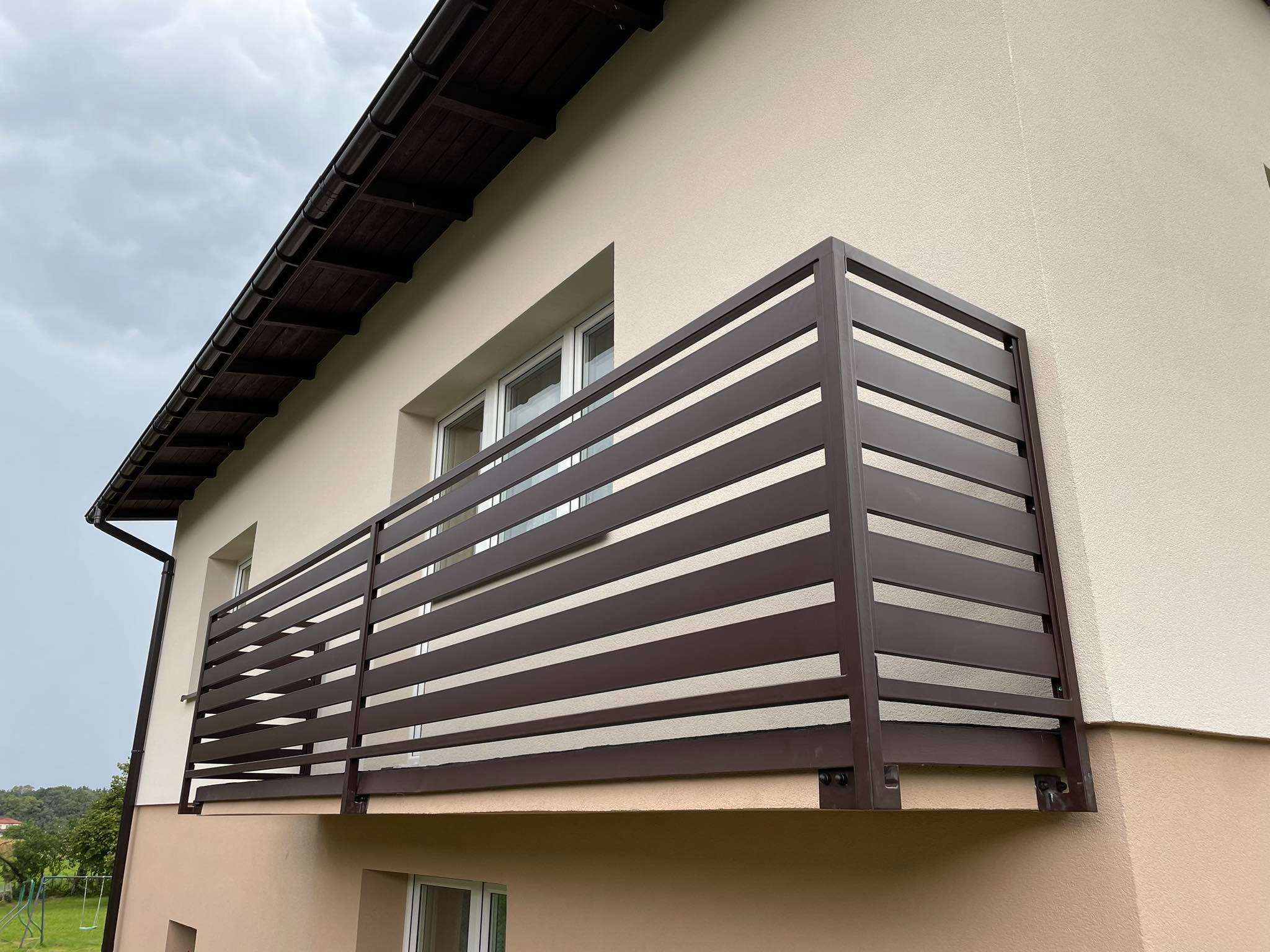 Nowoczesna, brązowa balustrada balkonowa z poziomych profili, zamontowana na elewacji domu w odcieniach beżu, widok z dołu na pochmurne niebo.