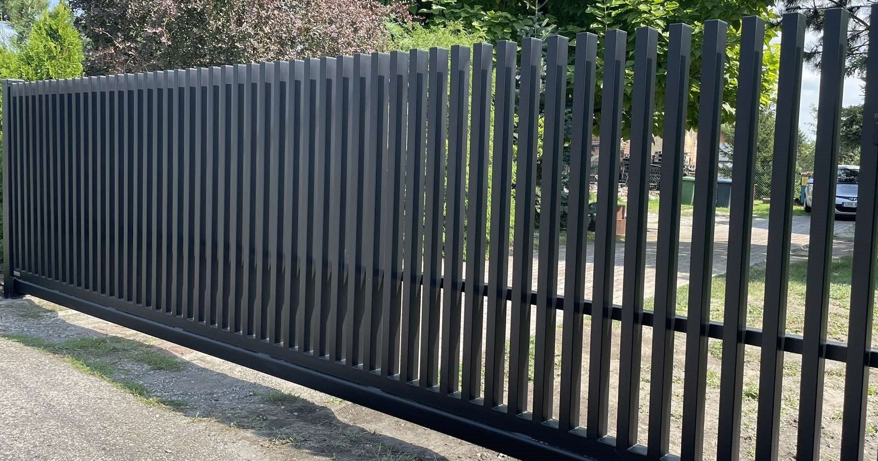 Nowoczesna, przesuwna brama wjazdowa w kolorze grafitowym z pionowymi profilami, widoczna z zewnątrz posesji, z zielenią w tle.