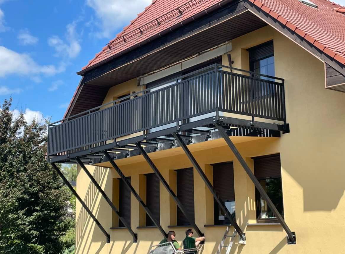 Montaż balkonu z czarną, metalową balustradą na żółtej elewacji budynku, widoczne ukośne wsporniki i dwóch monterów na podnośniku.