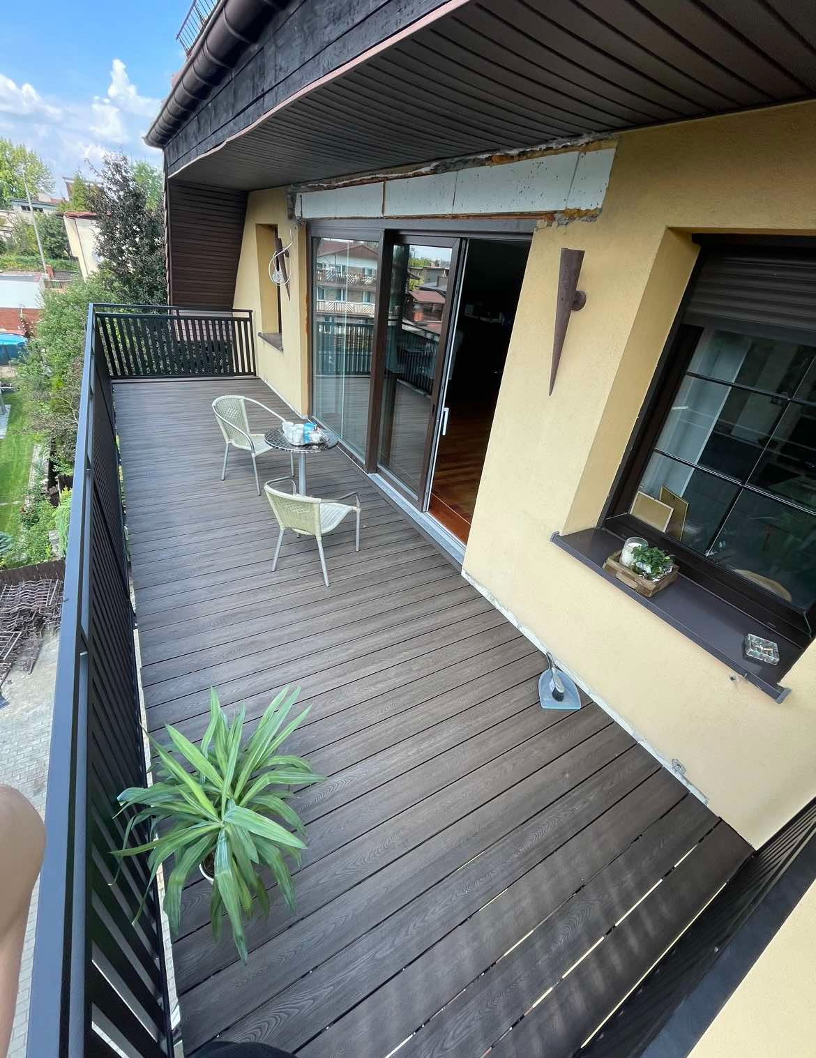 Zadaszony balkon z drewnopodobną podłogą, czarną balustradą z metalowych prętów, dwoma krzesłami i stolikiem kawowym, widok z góry, w tle zieleń.