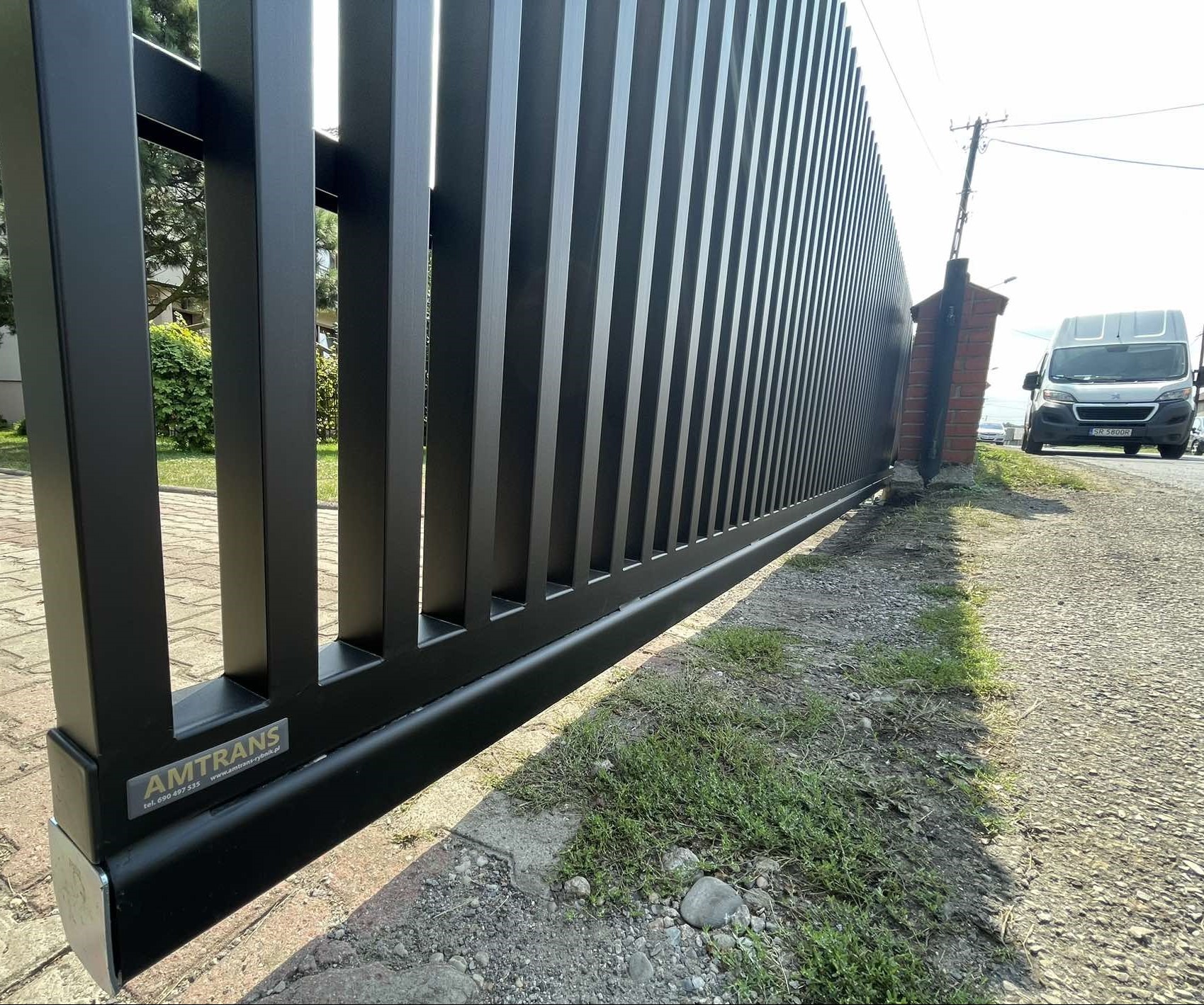 Nowoczesna, przesuwna brama wjazdowa z czarnymi pionowymi profilami, widoczne logo firmy AMTRANS na dolnej ramie, na tle kostka brukowa, trawa i zaparkowany biały bus.