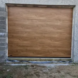 Zamknięta, segmentowa brama garażowa w kolorze jasnego drewna, zamontowana w surowym otworze budynku. Widoczne detale montażu i otoczenie placu budowy.