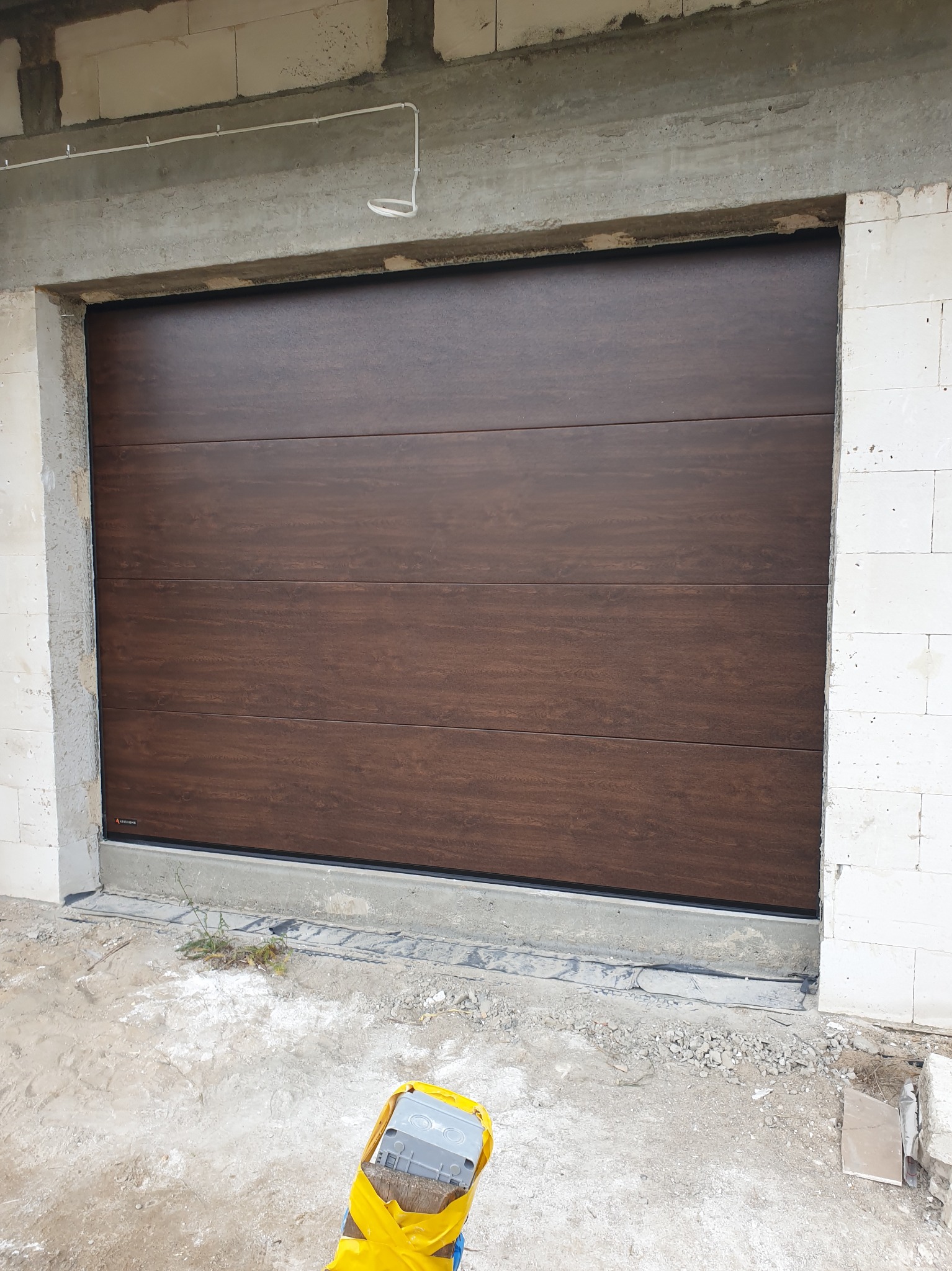 Zamknięta, brązowa brama garażowa z poziomymi panelami, zamontowana w surowym otworze budynku z białych bloczków. Widoczne elementy montażowe i fragment podłoża.
