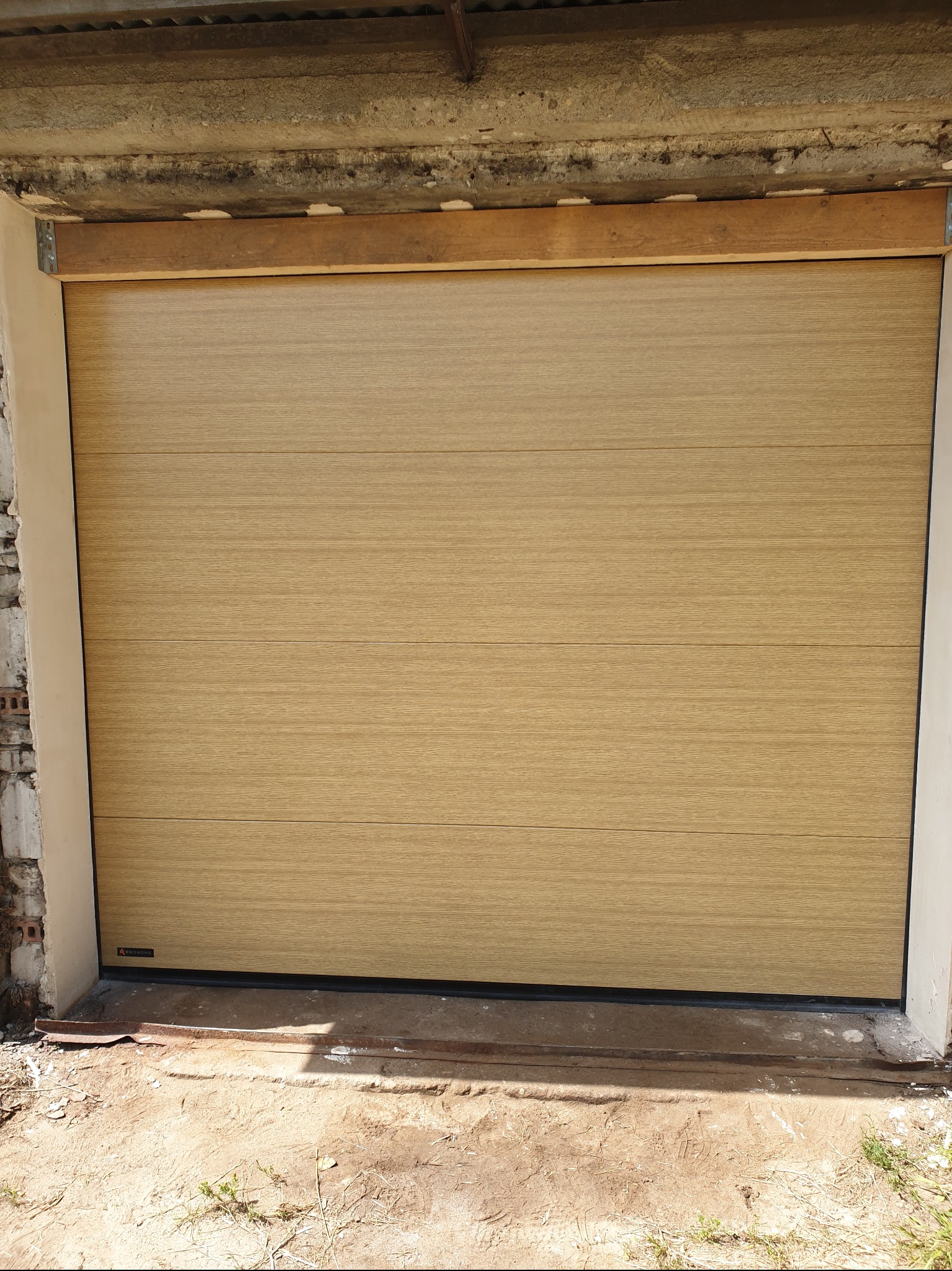 Jasna, segmentowa brama garażowa z poziomymi panelami o strukturze drewna. Widoczne mocowania i surowy beton nad bramą. Brama zamknięta, z logo producenta w dolnym rogu.