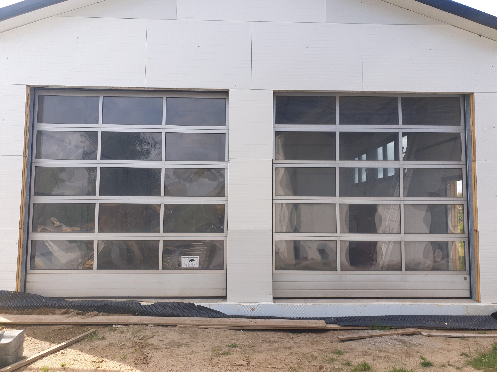 Dwie segmentowe bramy garażowe z przeszkleniami, zamontowane w białym budynku z paneli. Widoczne elementy konstrukcyjne i podłoże przed budynkiem.