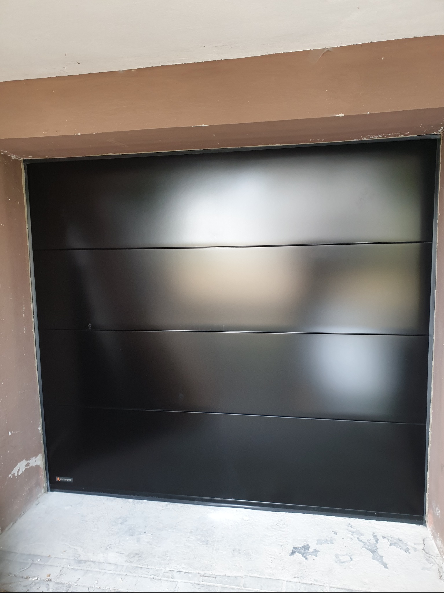 Czarna, segmentowa brama garażowa z poziomymi panelami, zamontowana w surowym, wykończonym wnęką garażową. Widoczne niedoskonałości ścian i podłoża.