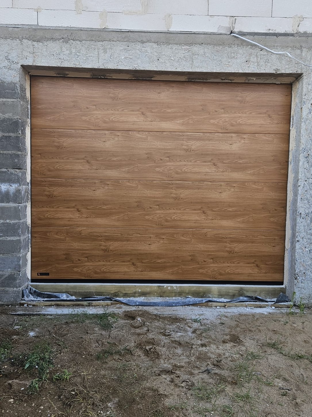 Zamknięta, segmentowa brama garażowa w kolorze jasnego drewna, zamontowana w surowym otworze budynku. Widoczne detale montażu i otoczenie placu budowy.