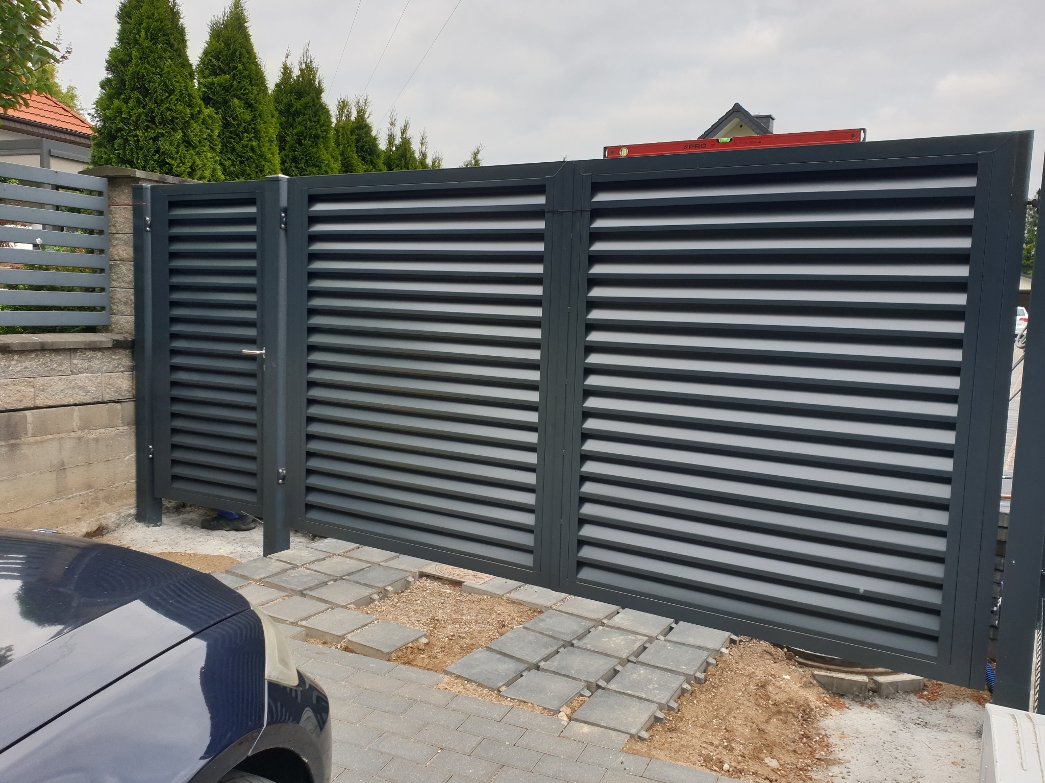 Nowoczesna, przesuwna, antracytowa brama z poziomymi panelami, widoczna od strony podjazdu. Częściowo ułożona kostka brukowa wokół bramy.