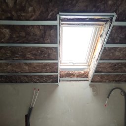 Mat Bet - Montaż izolacji z wełny mineralnej w poddaszu z oknem dachowym, widoczne profile metalowe i wystające rury instalacyjne.