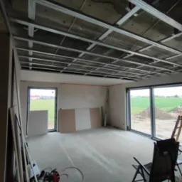 Wnętrze w trakcie montażu sufitu podwieszanego z płyt karton-gips, widoczne metalowe profile konstrukcyjne, płyty gipsowe opierające się o ściany, rozłożone narzędzia i krzesło turystyczne.