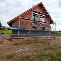 Mat Bet - Budowa domu jednorodzinnego: widok na niewykończoną elewację z czerwonej cegły i szarego ocieplenia, otoczoną metalowym rusztowaniem na tle zachmurzonego nieba.