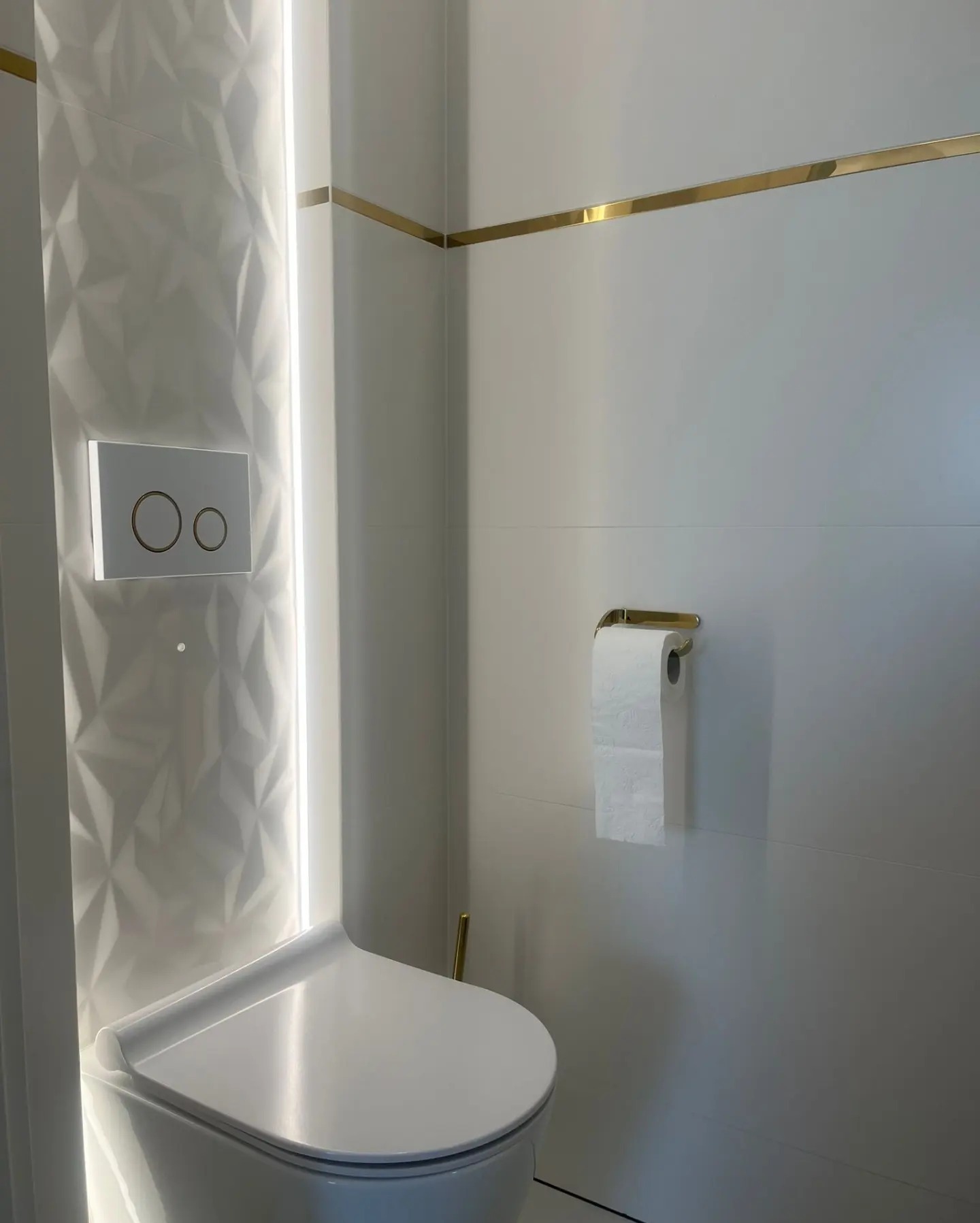 Nowoczesna toaleta w białej łazience z podświetlaną ścianą o geometrycznym wzorze i złotymi akcentami, takimi jak listwy i uchwyt na papier toaletowy.