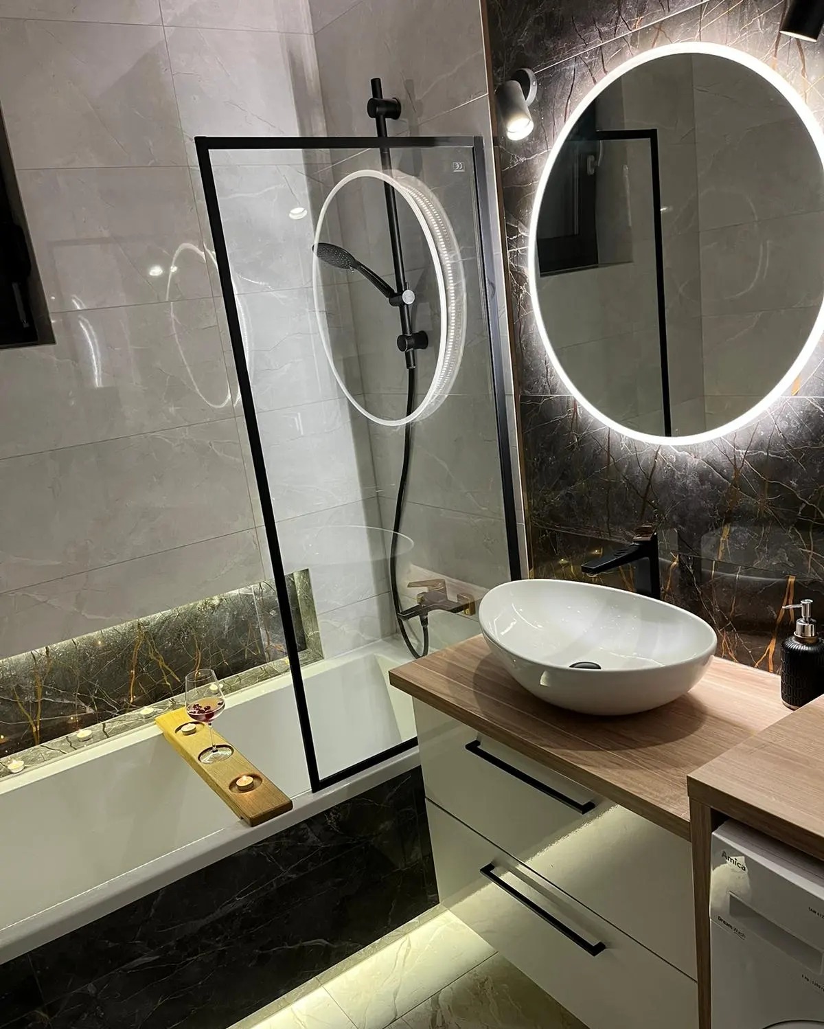 Nowoczesna łazienka z marmurowymi płytkami, kabiną prysznicową z czarnymi ramami, okrągłym lustrem z podświetleniem LED, białą umywalką nablatową na szafce z drewnianym blatem i wanną z drewnianą...