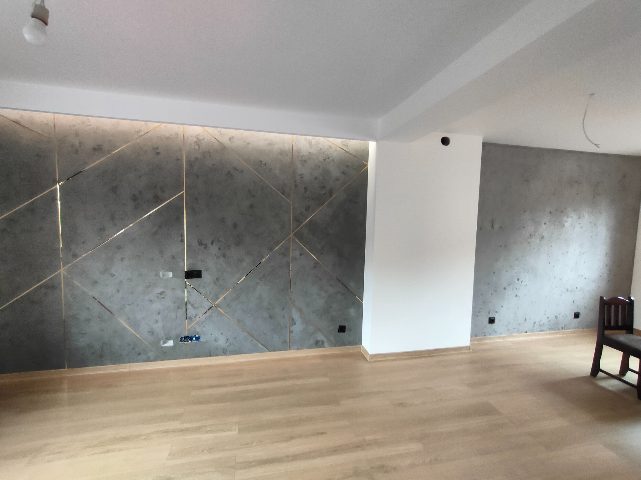 Nowoczesne wykończenie wnętrza z szarą ścianą dekoracyjną z geometrycznym wzorem w złotych liniach, jasna podłoga i białe ściany, widoczne gniazdka elektryczne.