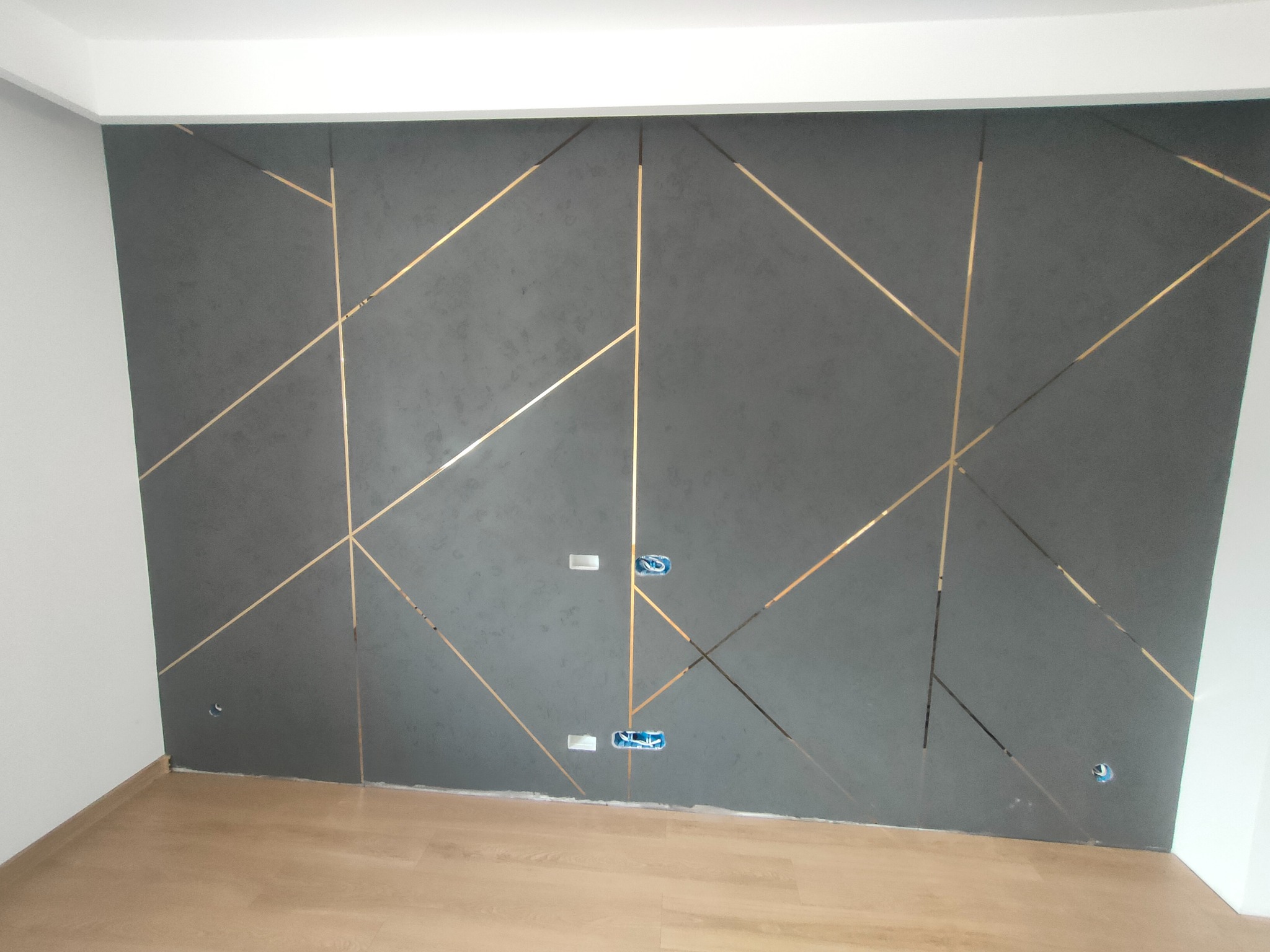 Szara ściana z dekoracyjnymi, złotymi paskami tworzącymi geometryczny wzór, widoczne gniazdka elektryczne bez osprzętu, jasna podłoga.