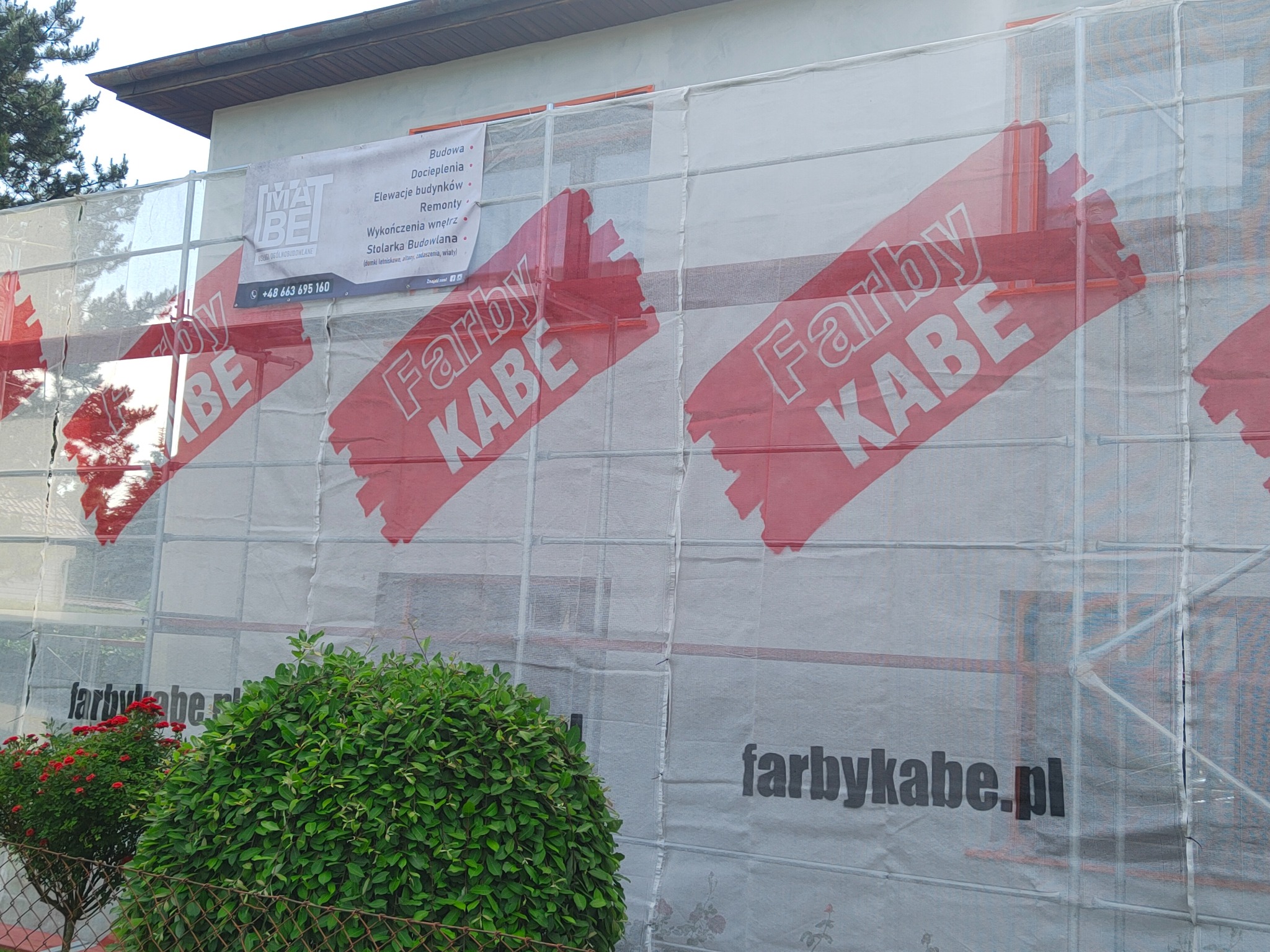 Budynek w trakcie prac remontowych, osłonięty siatką z reklamą farb KABE, widoczne rusztowania, krzewy i drzewa w tle.