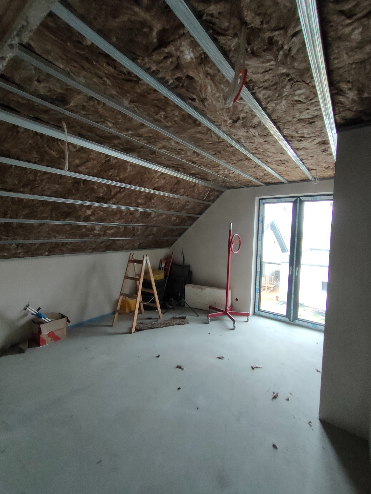 Poddasze w trakcie adaptacji, widoczna izolacja z wełny mineralnej między metalowymi profilami sufitowymi, drabina drewniana, podnośnik do płyt gipsowo-kartonowych i karton na podłodze.