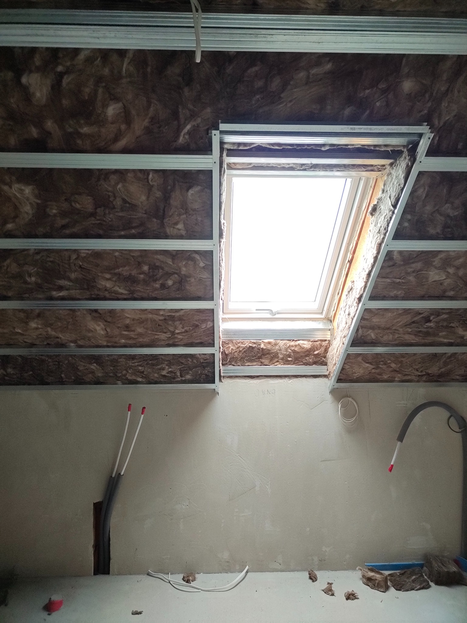 Montaż izolacji z wełny mineralnej w poddaszu z oknem dachowym, widoczne profile metalowe i wystające rury instalacyjne.