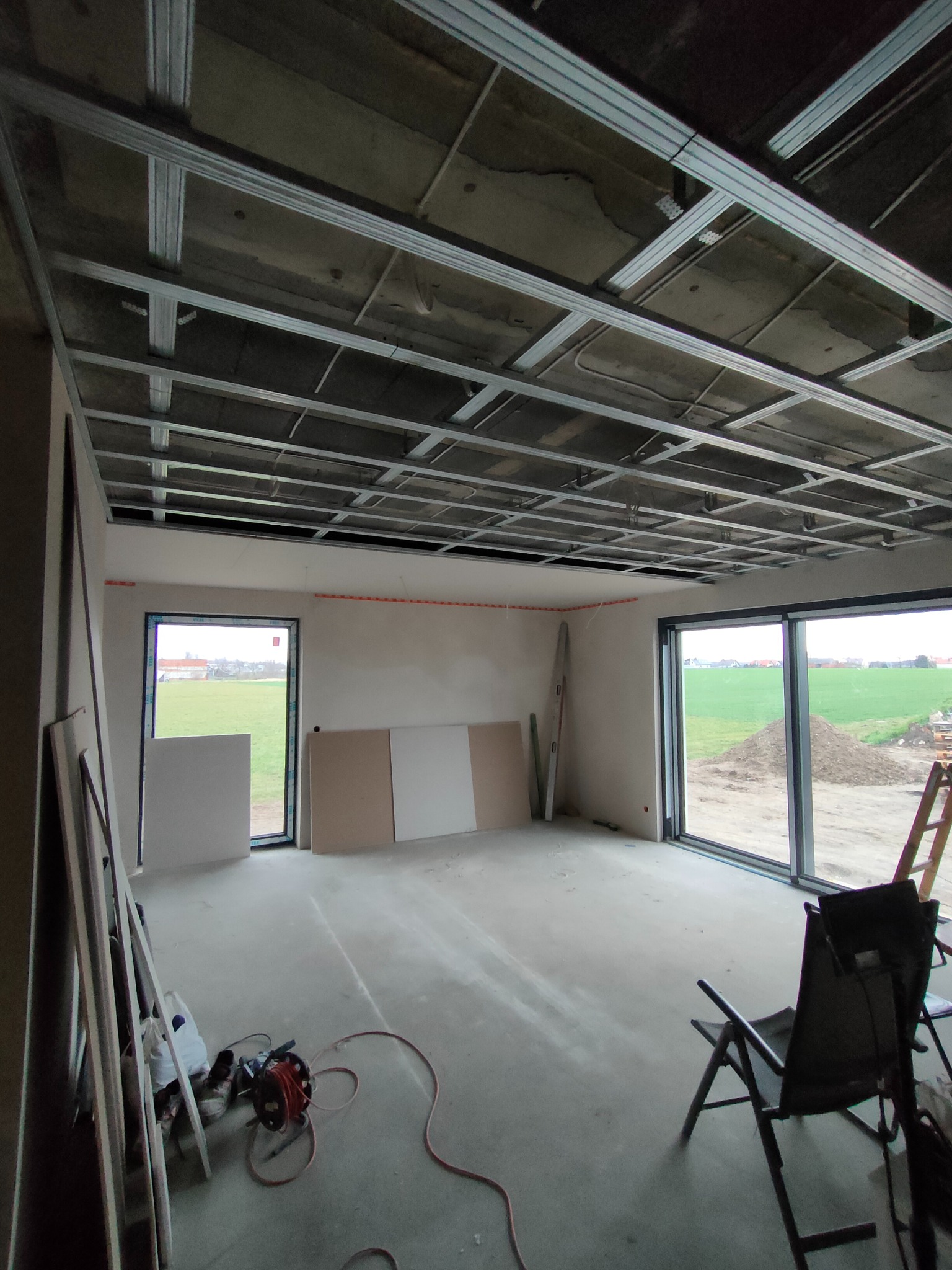 Wnętrze w trakcie montażu sufitu podwieszanego z płyt karton-gips, widoczne metalowe profile konstrukcyjne, płyty gipsowe opierające się o ściany, rozłożone narzędzia i krzesło turystyczne.