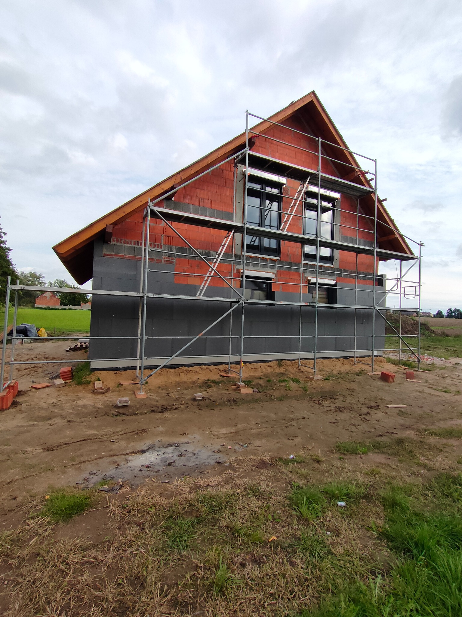Budowa domu jednorodzinnego: widok na niewykończoną elewację z czerwonej cegły i szarego ocieplenia, otoczoną metalowym rusztowaniem na tle zachmurzonego nieba.