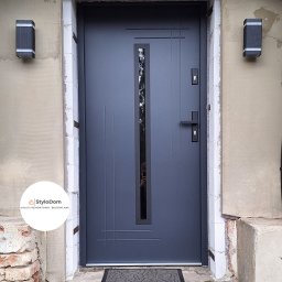 StyloDom Igor Pająk - Szare drzwi wejściowe z szybą, zamontowane w ościeżnicy z widocznymi śladami remontu, oświetlone dwoma zewnętrznymi lampami, z wycieraczką przed progiem i logo firmy remontowej w lewym dolnym rogu.