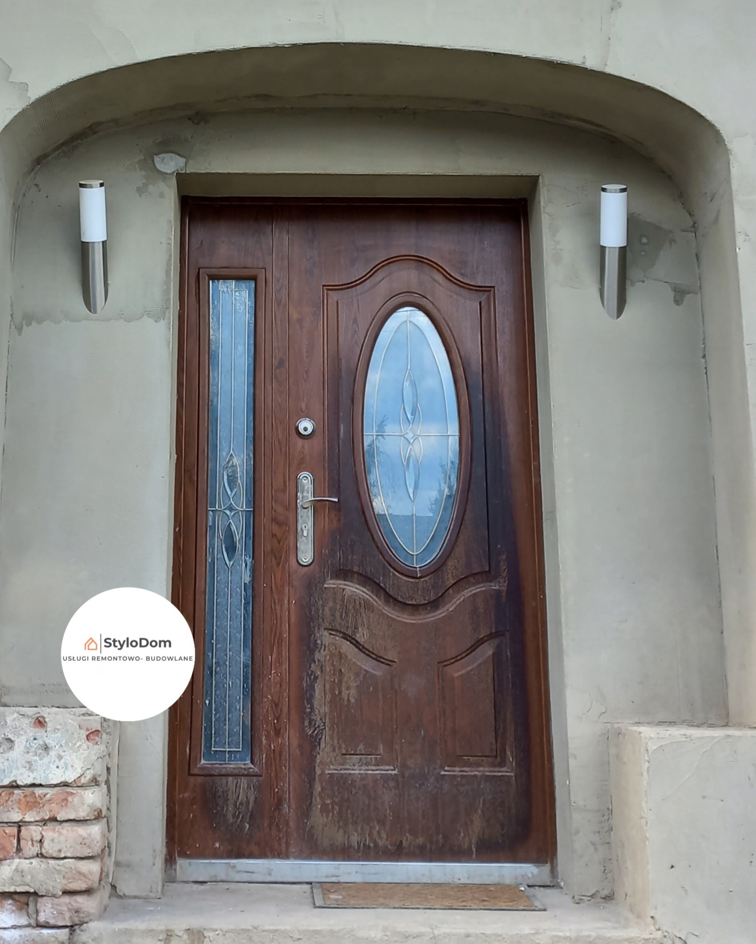 Ciemnobrązowe drzwi wejściowe z owalnym i prostokątnym przeszkleniem, zamontowane w surowym otworze z widocznymi śladami tynku i cegłami u podstawy, oświetlone dwoma pionowymi lampami ściennymi po...