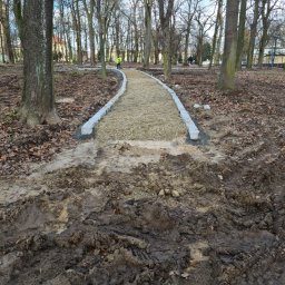 J&M SPÓŁKA Z OGRANICZONĄ ODPOWIEDZIALNOŚCIĄ - Budowa ścieżki w parku: ułożone obrzeża z kostki granitowej i wysypany żwir, w tle drzewa i pracownik w kamizelce odblaskowej, w pierwszym planie błoto.