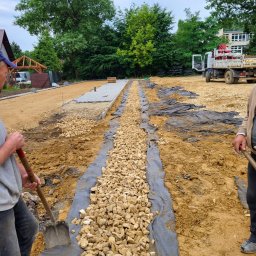 J&M SPÓŁKA Z OGRANICZONĄ ODPOWIEDZIALNOŚCIĄ - Przygotowanie podłoża pod ścieżkę z kostki: warstwa drenażowa z kamienia na geowłókninie, widoczni pracownicy z łopatami, w tle ułożona część kostki i ciężarówka