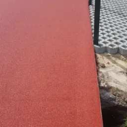 Czerwona, gumowa nawierzchnia boiska sportowego przylegająca do ażurowych, betonowych płyt chodnikowych i zielonych słupk&oacute;w ogrodzenia.