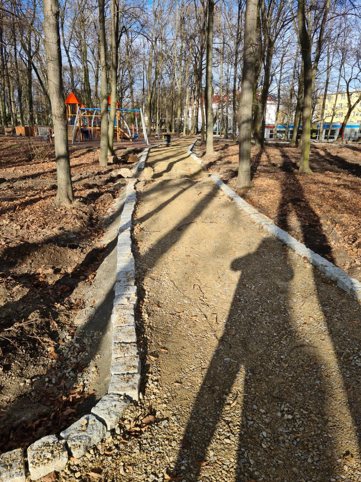 Ścieżka w parku w trakcie budowy, z obrzeżami z kostki granitowej, widoczne drzewa i plac zabaw w tle, cień osoby fotografującej na ścieżce.