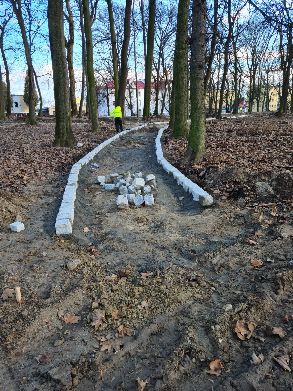 Budowa ścieżki w parku: ułożone obrzeża z jasnoszarej kostki granitowej, przygotowane miejsce pod nawierzchnię, w tle pracownik w żółtej kamizelce i drzewa.
