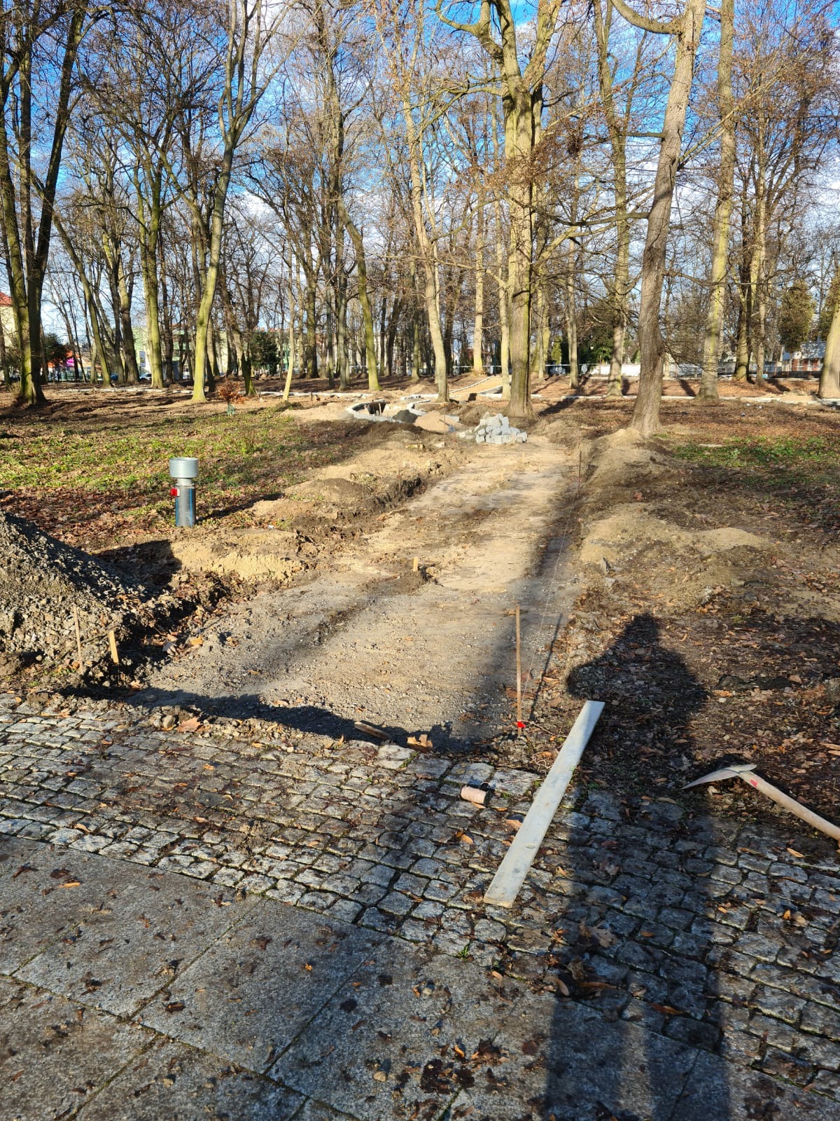 Przygotowanie podłoża pod ścieżkę w parku, widoczna wykopana ziemia, obrzeża, paliki wyznaczające trasę, narzędzia i cień osoby wykonującej prace.