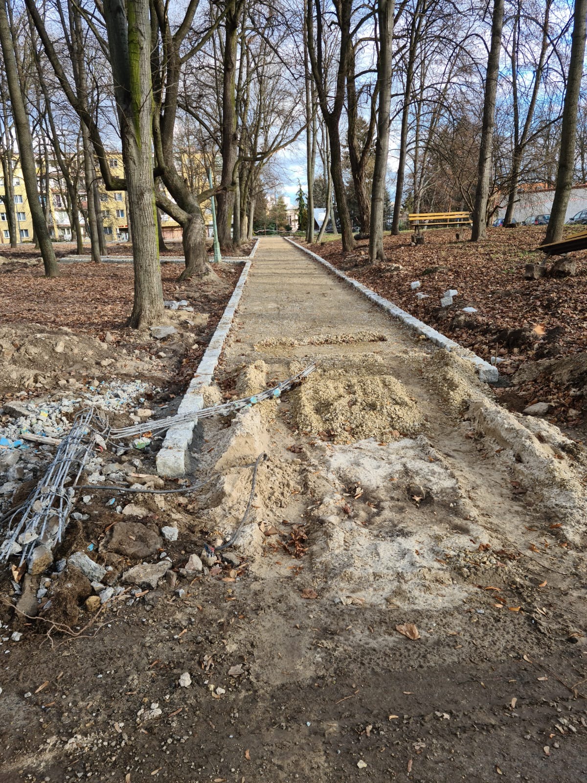 Budowa ścieżki w parku z obrzeżami z jasnej kostki, widoczne przygotowane podłoże i materiały budowlane, drzewa w tle.