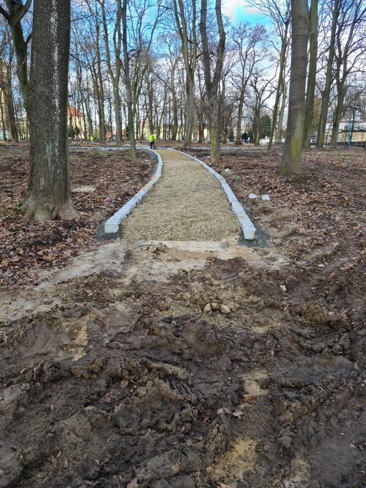 Budowa ścieżki w parku: ułożone obrzeża z kostki granitowej i wysypany żwir, w tle drzewa i pracownik w kamizelce odblaskowej, w pierwszym planie błoto.