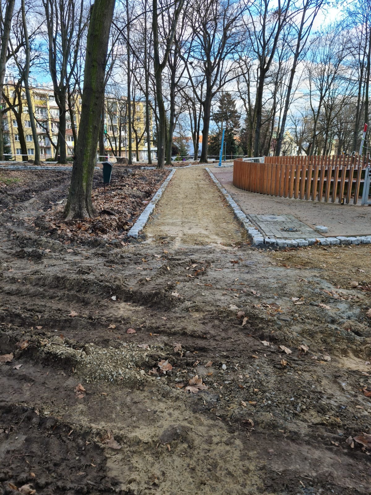 Ścieżka w parku w trakcie budowy, obrzeża z kostki granitowej, widoczne ślady po maszynach budowlanych na ziemi, w tle drzewa i budynek.