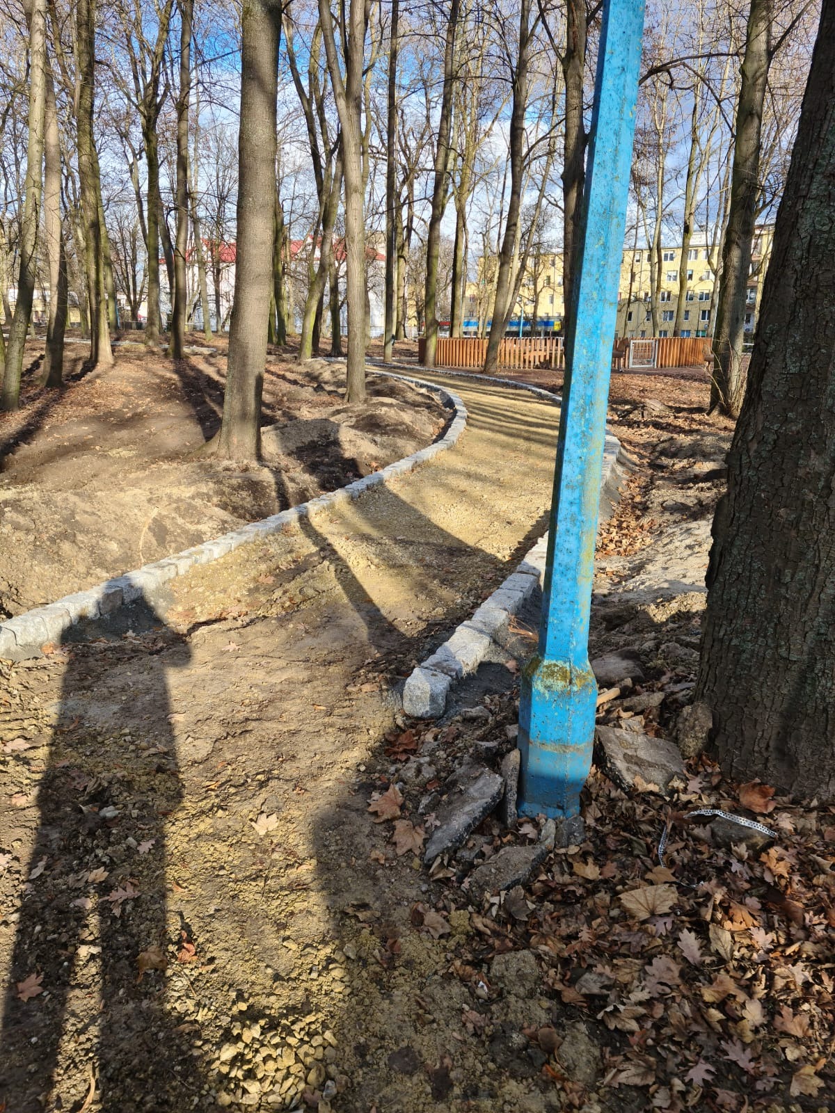Budowa ścieżki w parku: ułożone obrzeża z kostki granitowej, podsypka z kruszywa, w tle drzewa i budynek, rzucający się w oczy niebieski słup
