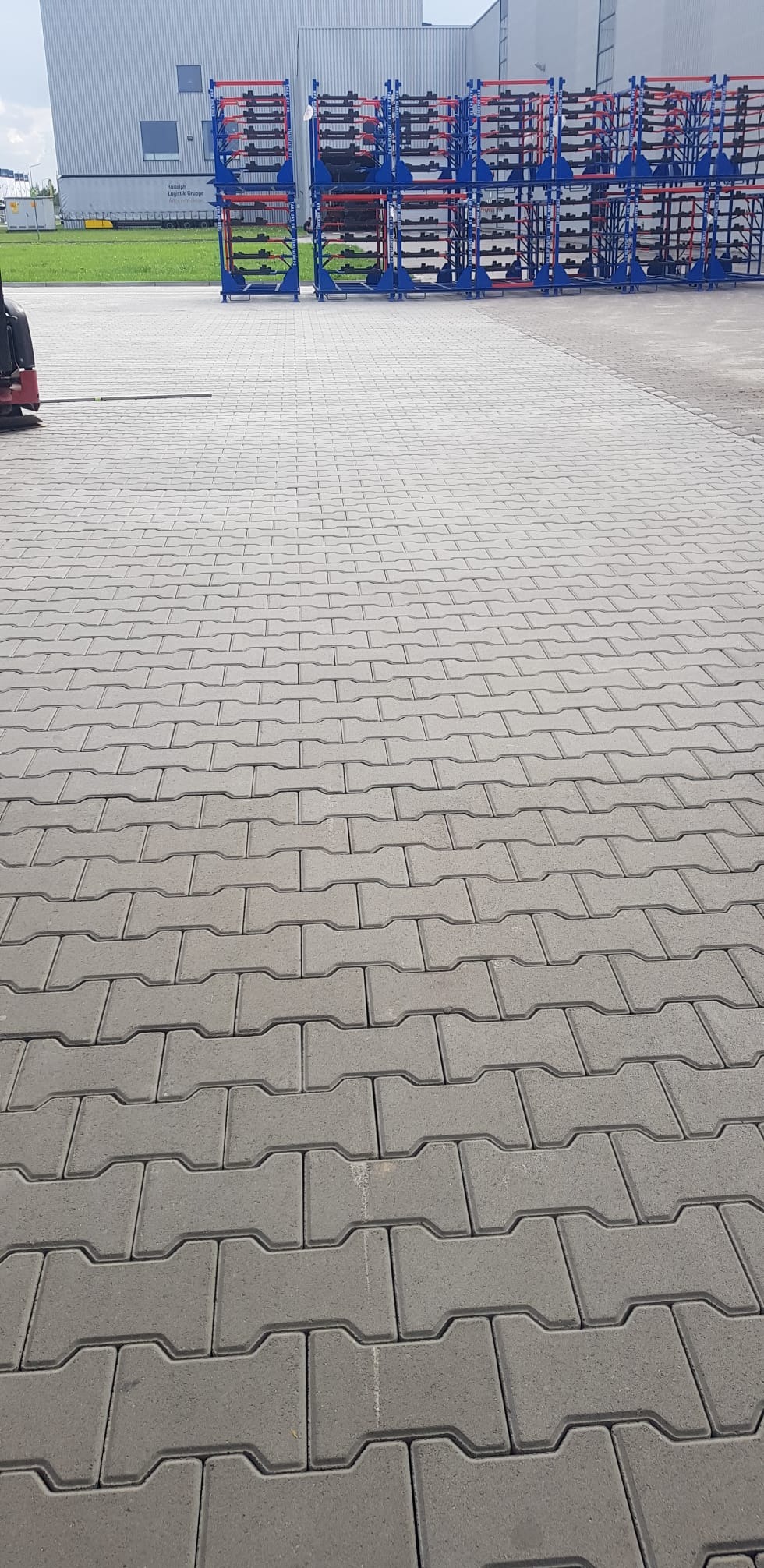 Szara kostka brukowa ułożona na parkingu przemysłowym, w tle regały magazynowe i budynek hali.