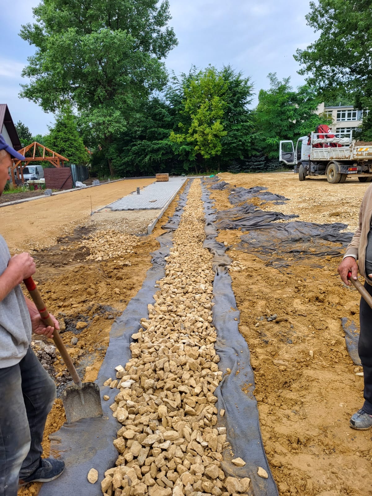 Przygotowanie podłoża pod ścieżkę z kostki: warstwa drenażowa z kamienia na geowłókninie, widoczni pracownicy z łopatami, w tle ułożona część kostki i ciężarówka