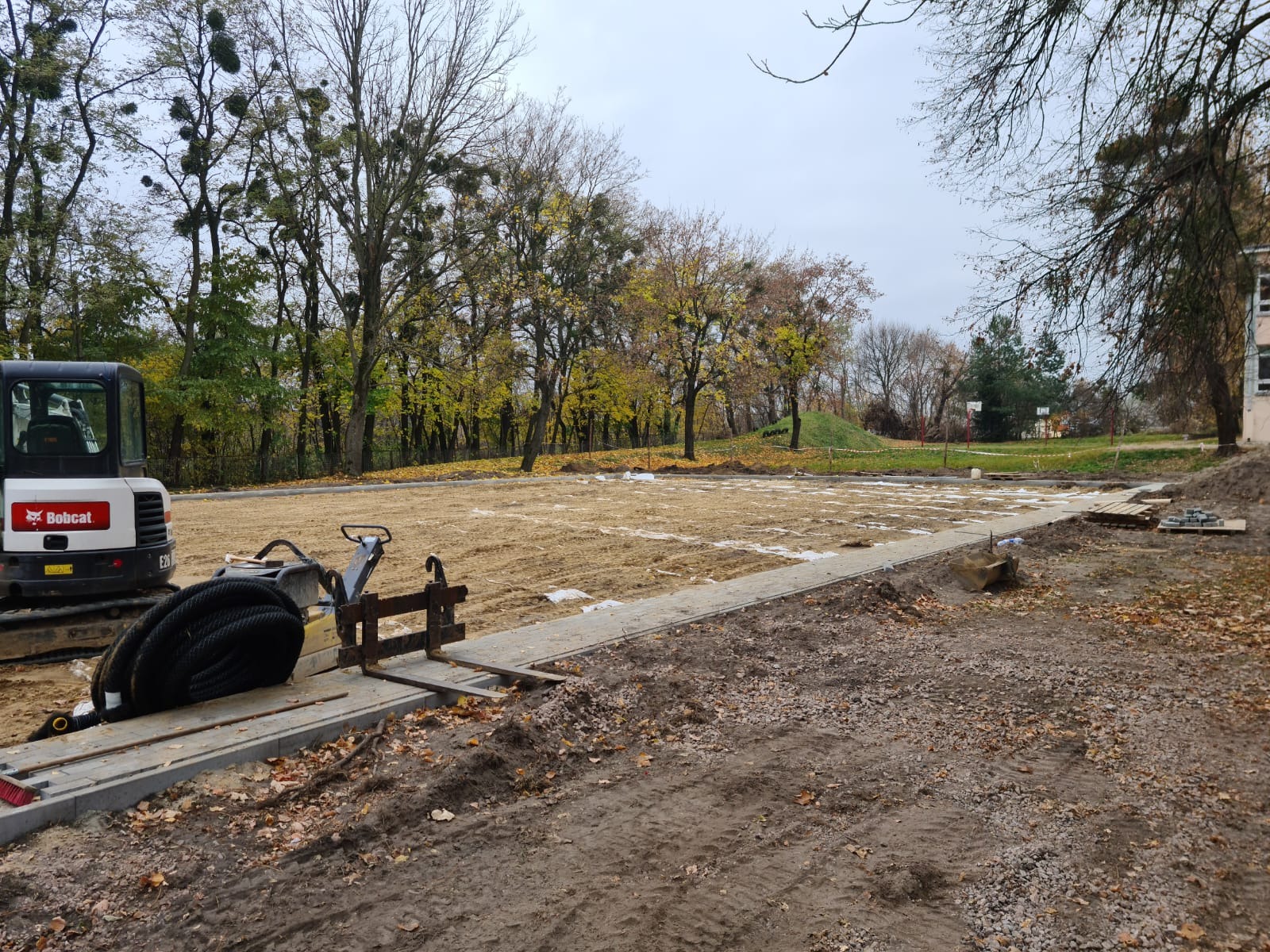 Przygotowanie terenu pod budowę placu: wyrównany piasek z widocznym zarysem przyszłej nawierzchni, obok minikoparka Bobcat i narzędzia, jesienne drzewa w tle.