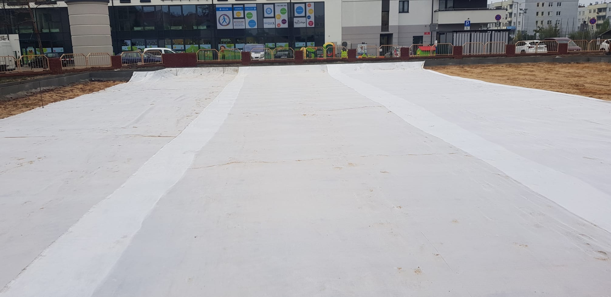 Przygotowanie podłoża pod budowę, teren wyłożony białą geowłókniną, widoczne obrzeża i budynki w tle.