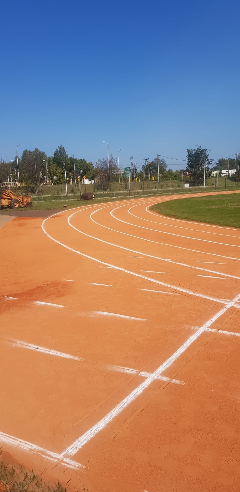 Pomarańczowa bieżnia lekkoatletyczna z białymi liniami torów, widziana pod kątem z bliska, z widocznym fragmentem traktora w tle i błękitnym niebem.