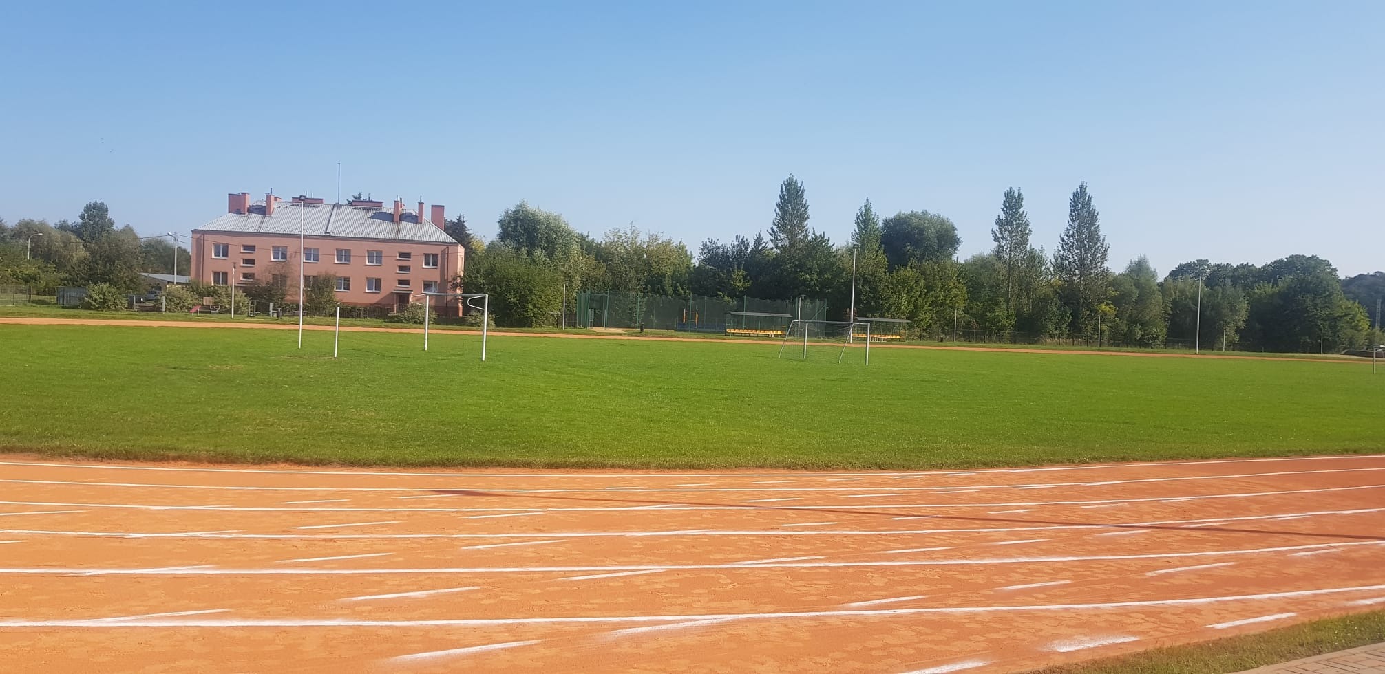 Widok na bieżnię lekkoatletyczną z pomarańczową nawierzchnią, zielone boisko i budynek w tle w słoneczny dzień.