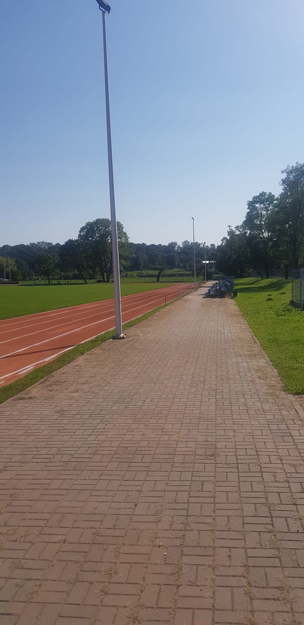 Ceglana ścieżka prowadząca wzdłuż bieżni lekkoatletycznej, oświetlona przez wysokie latarnie, z zielonym trawnikiem i drzewami w tle, przy słonecznej pogodzie.