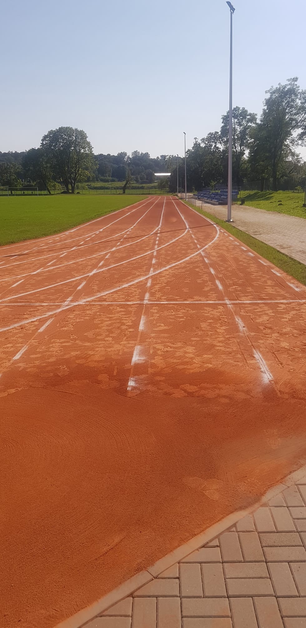 Pomarańczowa bieżnia lekkoatletyczna z białymi liniami torów, widok perspektywiczny w kierunku trybun i oświetlenia, obrzeże z kostki brukowej.