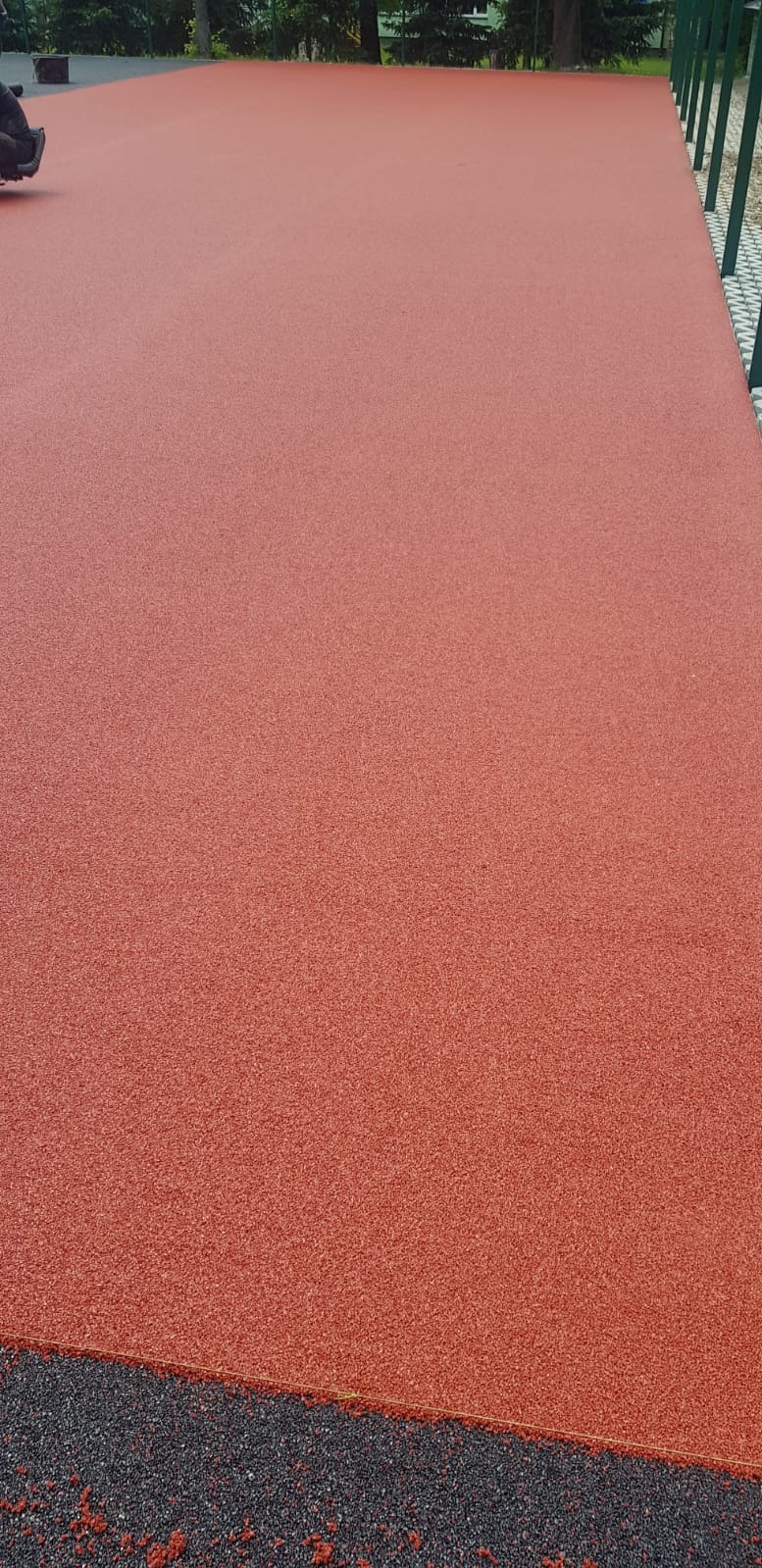 Czerwona, gumowa nawierzchnia boiska sportowego lub placu zabaw, widoczne czarne obrzeże i fragment zielonego ogrodzenia.